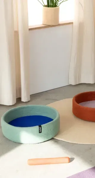 Camas para perros hechas a mano y sostenibles en fieltro azul cielo con interior azul real y terracota con interior beige, colocadas sobre alfombras en tonos neutros. Juguetes de fieltro en tonos cálidos a su alrededor.