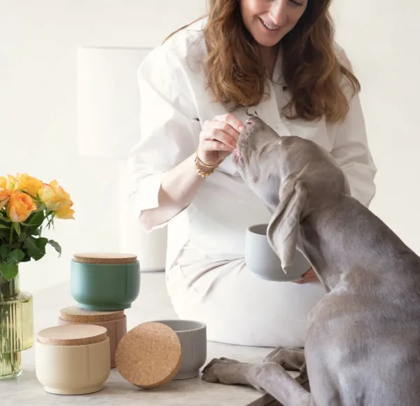 Una mujer sonriente vestida de blanco ofrece un premio a su perro de gran tamaño, que se apoya en la encimera para alcanzarlo. Ella sostiene uno de los frascos de cerámica gris claro con tapa de corcho, mientras que sobre la mesa hay otros tres botes en colores beige, rosa y verde. El espacio está decorado con flores amarillas en un jarrón y una lámpara de fondo. La imagen transmite cercanía, cariño y bienestar, mostrando cómo estos recipientes pueden integrarse en una rutina diaria con estilo y funcionalidad.