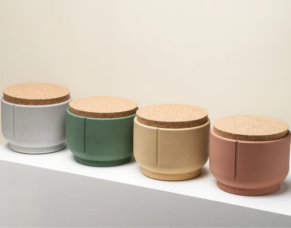 Cuatro recipientes artesanales para snacks de perro están alineados sobre una superficie blanca, con un fondo liso beige. Los envases están hechos a mano en cerámica y muestran una gama de colores naturales: gris claro, verde oscuro, arena cálida y terracota. Cada uno tiene una tapa de corcho gruesa que sella el contenido de forma hermética. La luz suave resalta la textura mate y elegante de los materiales. Es una escena limpia y minimalista que transmite calma y diseño consciente.