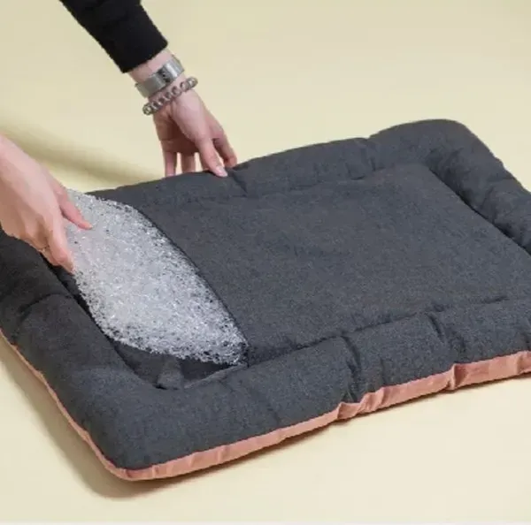 Cama ortopédica para perros en tono gris oscuro con bordes rosa, mostrando el relleno transparente y transpirable. Una persona coloca el material en la funda, destacando la facilidad de limpieza y el soporte ergonómico para un descanso cómodo y saludable.