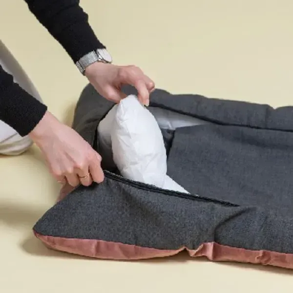 Cama ortopédica para perros de color gris oscuro y borde rosa, mostrando cómo se coloca el relleno blanco dentro de la funda. Una persona cierra la cremallera, destacando la practicidad, la limpieza sencilla y el confort ortopédico para los perros.